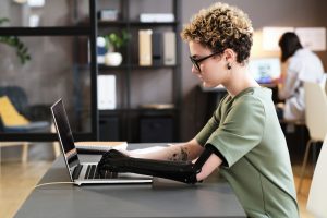 Young woman with prosthetic arm typing on laptop while sitting at office desk By AnnaStills